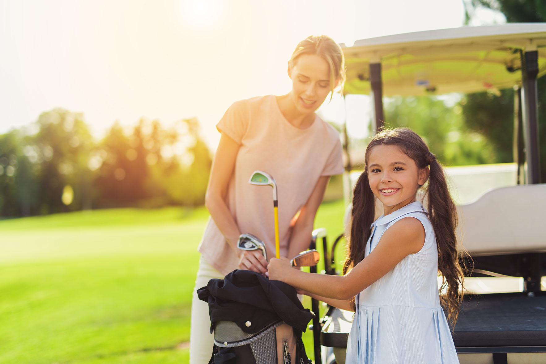 The girl takes out the golf club from the bag and looks into the lens. A woman is standing behind her and looking at the golf club she is pulling out