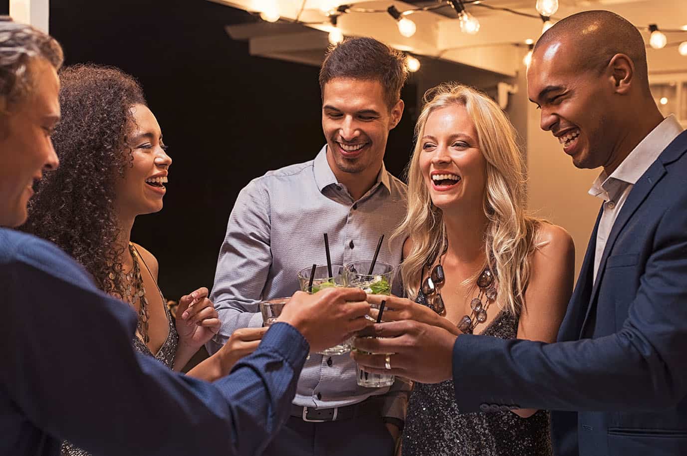 Group of young multiethnic friends enjoying evening and drinking cocktails. Happy men and women raising a toast with mojito on a patio under the light bulb wire. Elegant girls and stylish guy having fun together at party night.